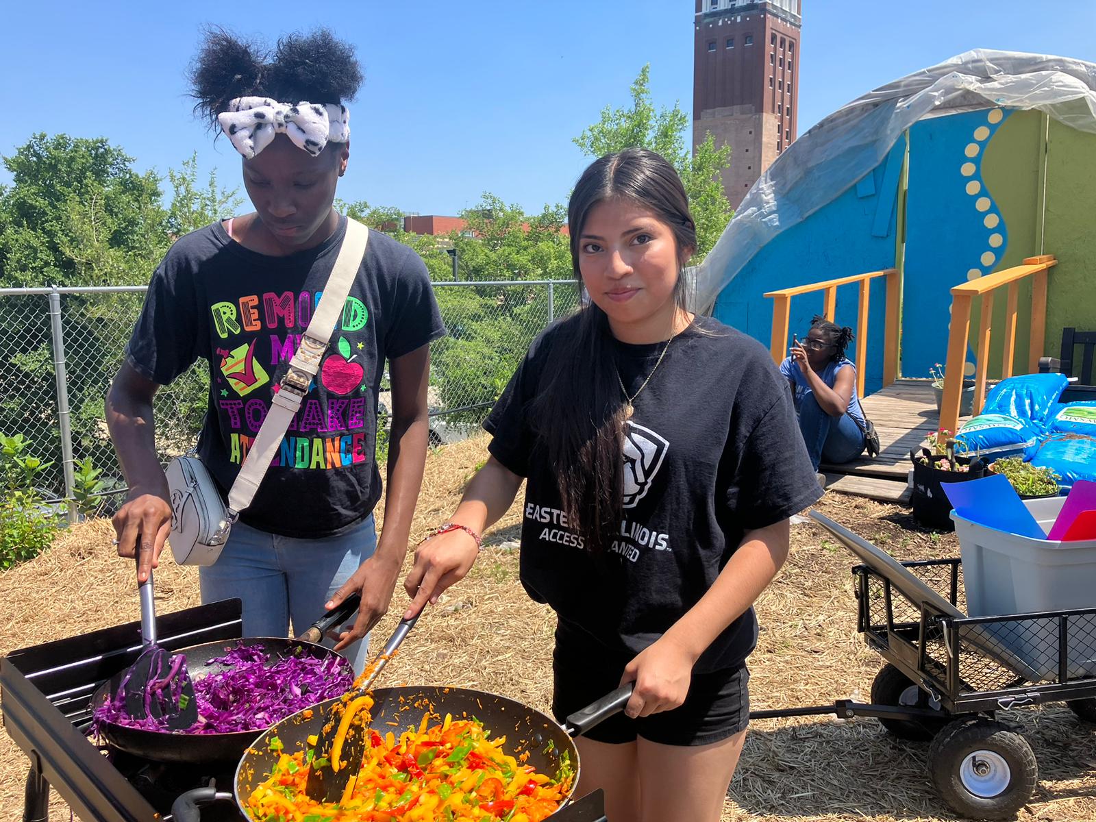 Two students cook veggies on an outdoor stove